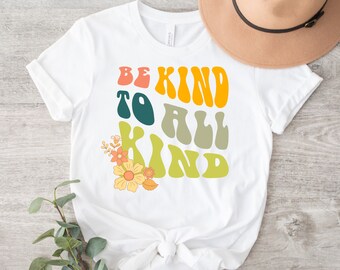 Pode incluir: Camiseta branca com um gráfico de estilo retro que diz "Be Kind to All Kind" em letras coloridas e onduladas. O gráfico é rodeado por um design floral.