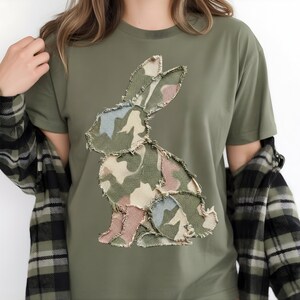 May include: An olive green t-shirt with a camouflage bunny applique. The bunny design uses green, beige, pink, and blue tones. The shirt has short sleeves and a round neckline.