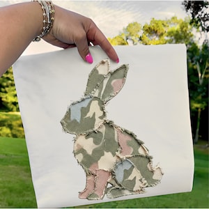 May include: A white canvas featuring a patchwork bunny design. The bunny is made of camouflage fabric in shades of green, pink, and blue. The edges of the fabric are frayed, giving it a rustic look. The design is held by a hand with pink nail polish.