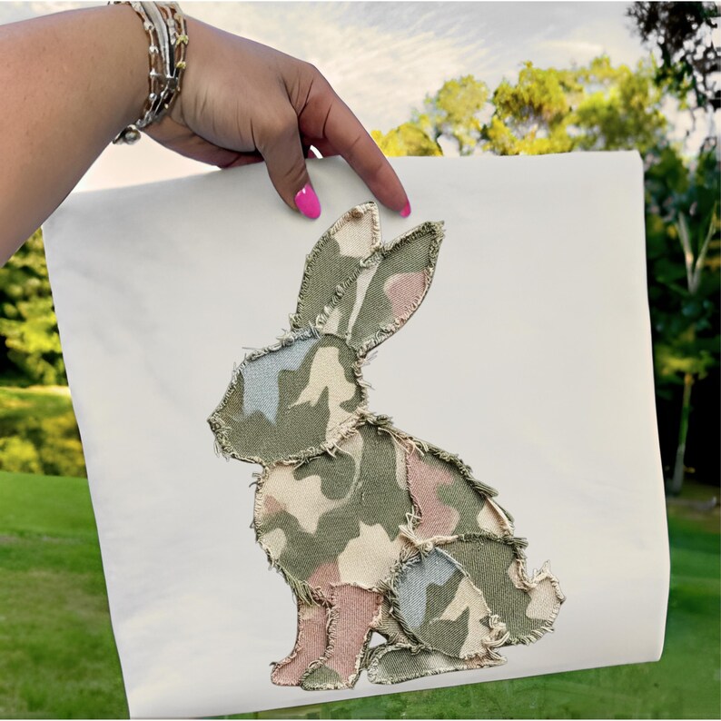 May include: A white canvas with a patchwork bunny design. The bunny is made of camouflage fabric in shades of green, pink, and blue. The fabric edges are frayed, giving a rustic look. The design is held by a hand with pink nail polish.