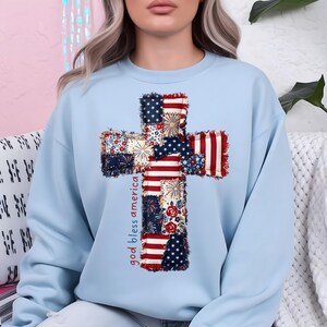 May include: A light blue sweatshirt featuring a cross design made of patchwork fabric with red, white, and blue patterns. The words "god bless america" are printed vertically on the left side of the cross.