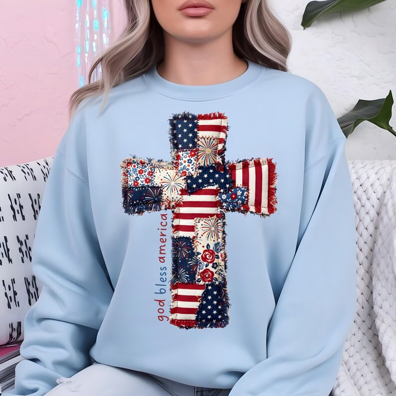 May include: A light blue sweatshirt featuring a cross design made of patchwork fabric with red, white, and blue patterns. The words "god bless america" are printed vertically on the left side of the cross.