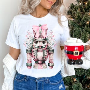 May include: White t-shirt featuring a watercolour illustration of a nutcracker in shades of pink and green. The nutcracker has a pink bow on its hat and a moustache.