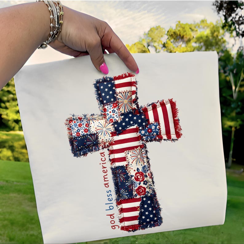 May include: A white fabric with a cross design featuring red, white, and blue patchwork patterns. The cross incorporates elements of the American flag, including stars and stripes. The words "god bless america" are printed in red.