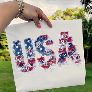 Puede incluir: Una tela color crema con las letras "USA" aplicadas en tela roja, blanca y azul. Las letras están decoradas con estampados florales y estrellas, con un borde deshilachado.