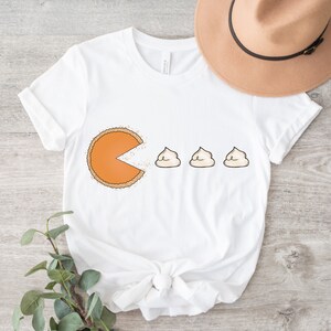 Puede incluir: Camiseta blanca con un gr&aacute;fico de una rebanada de pastel de calabaza con la boca abierta, comiendo tres peque&ntilde;os malvaviscos blancos. El texto "Pac-Pie" est&aacute; impreso en la camiseta.