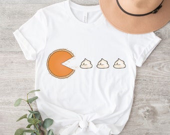 May include: White t-shirt with a graphic of a pumpkin pie slice with an open mouth, eating three small white marshmallows. The text "Pac-Pie" is printed on the shirt.