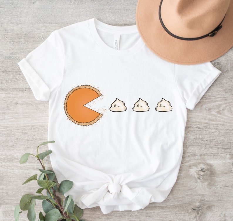 Puede incluir: Camiseta blanca con un gr&aacute;fico de una rebanada de pastel de calabaza con la boca abierta, comiendo tres peque&ntilde;os malvaviscos blancos. El texto "Pac-Pie" est&aacute; impreso en la camiseta.
