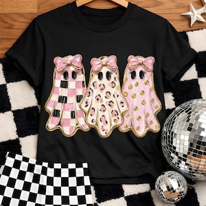 May include: A black short-sleeve t-shirt featuring a graphic of three ghosts with pink bows. The first ghost has a pink and black checkered pattern, the second has a leopard print, and the third has gold polka dots, all with textured paint effects.