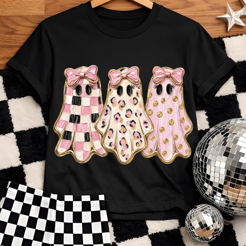 May include: A black short-sleeve t-shirt featuring a graphic of three ghosts with pink bows. The first ghost has a pink and black checkered pattern, the second has a leopard print, and the third has gold polka dots, all with textured paint effects.