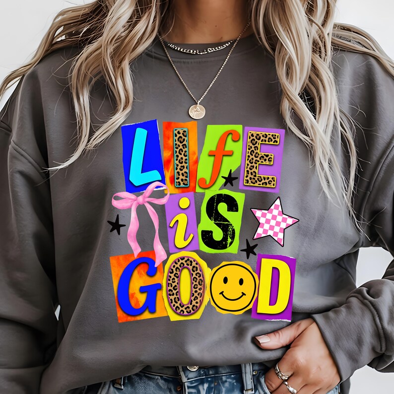 Może przedstawiać: Szara bluza z napisem "LIFE is GOOD" w kolorowych, blokowych literach. Litery mają r&oacute;żne wzory, w tym nadruk w cętki i emotikon uśmiechu. R&oacute;żowa kokarda i gwiazdki dopełniają projekt.