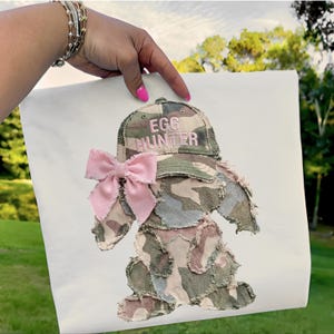May include: A white fabric with an appliqued Easter bunny design. The bunny is made of camouflage fabric with a pink bow and a hat that says "EGG HUNTER" in pink embroidery. The design is held up by a hand.