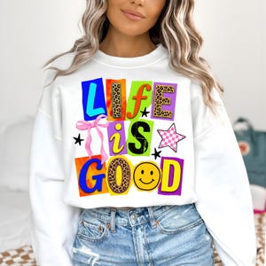 Może przedstawiać: Biała bluza z napisem "LIFE IS GOOD" w kolorowym, blokowym wzorze liter. Litery mają r&oacute;żne wzory, w tym nadruk w cętki i emotikon uśmiechu. R&oacute;żowa kokarda i gwiazdki dopełniają projekt.