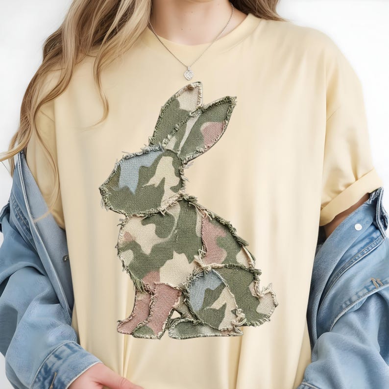 May include: A pale yellow t-shirt with a large appliqu&eacute; of a rabbit. The rabbit design is made from camouflage fabric in green, pink, and blue. The shirt has a relaxed fit and rolled-up sleeves, suggesting a casual style.