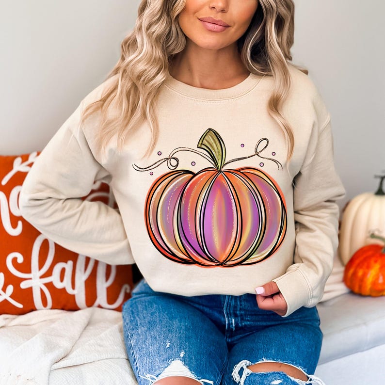 May include: A beige sweatshirt with a colorful pumpkin graphic. The pumpkin is outlined in black and has a swirl design. The sweatshirt has long sleeves and a crew neck.