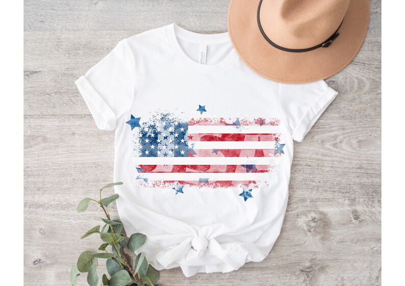 May include: White t-shirt with a red, white, and blue watercolor American flag design. The flag is distressed and has stars around it.
