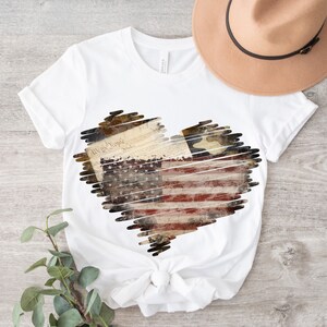 Puede incluir: Camiseta blanca con un dise&ntilde;o en forma de coraz&oacute;n que presenta una bandera estadounidense desgastada, la Declaraci&oacute;n de Independencia y una estrella. El dise&ntilde;o est&aacute; delineado con un efecto garabateado.
