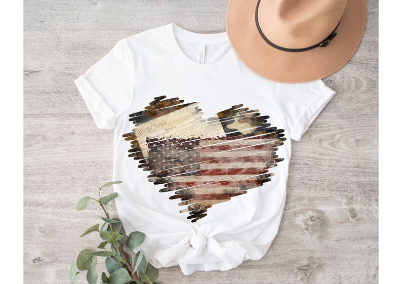 Puede incluir: Camiseta blanca con un dise&ntilde;o en forma de coraz&oacute;n que presenta una bandera estadounidense desgastada, la Declaraci&oacute;n de Independencia y una estrella. El dise&ntilde;o est&aacute; delineado con un efecto garabateado.