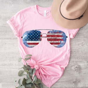 K&ouml;nnte beinhalten: Pinkes T-Shirt mit einem Aufdruck von Aviator-Sonnenbrillen mit amerikanischem Flaggenmuster.