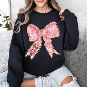May include: Black sweatshirt with a pink bow design featuring a nutcracker and other Christmas elements. The bow is decorated with gold glitter.