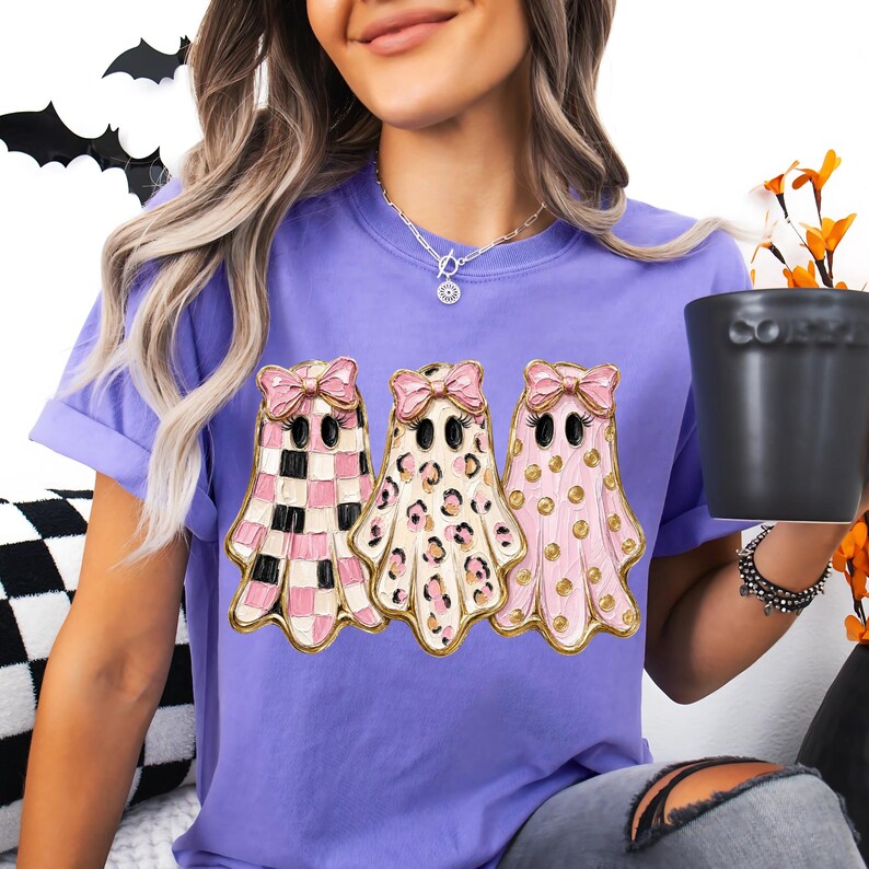May include: A purple t-shirt featuring a graphic of three ghosts, each wearing a pink bow. The first ghost has a pink and white checkered pattern, the second has a leopard print, and the third has pink polka dots on a light pink background.