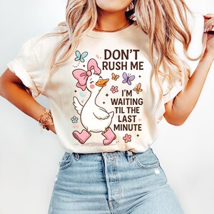 May include: A cream-coloured t-shirt with a cartoon goose wearing a pink bow and boots, and the text "DON'T RUSH ME I'M WAITING TIL THE LAST MINUTE." Butterflies and flowers decorate the design. The shirt is worn with blue jeans.