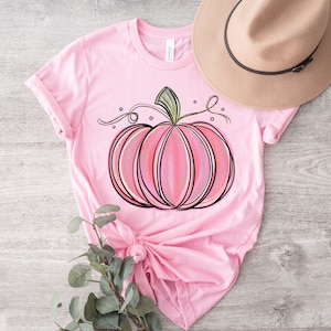 May include: A pink t-shirt with a pink and white pumpkin graphic. The pumpkin has a green stem and vine.