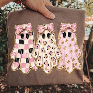 May include: A brown t-shirt featuring a graphic of three ghosts with pink bows and eyelashes. The first ghost has a pink and black checkerboard pattern, the second has a pink and black leopard print, and the third is pink with gold polka dots.