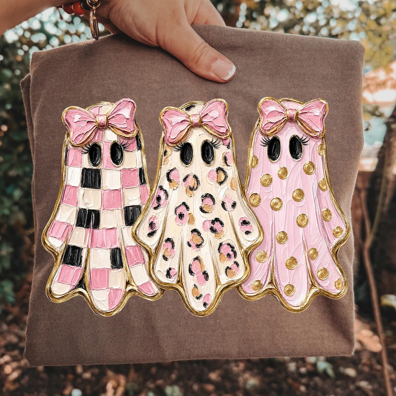 May include: A brown t-shirt featuring a graphic of three ghosts with pink bows and eyelashes. The first ghost has a pink and black checkerboard pattern, the second has a pink and black leopard print, and the third is pink with gold polka dots.