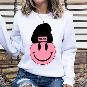 K&ouml;nnte beinhalten: Wei&szlig;er Pullover mit einem rosafarbenen Smiley, der eine schwarze M&uuml;tze mit dem Wort "MAMA" tr&auml;gt.
