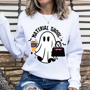 May include: White sweatshirt with a ghost graphic holding a coffee cup and a purse. The text "Material Ghoul" is written in a black circle around the ghost.