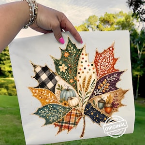May include: A white fabric square featuring a large maple leaf graphic divided into segments with various patterns including black and white buffalo plaid, orange paisley, polka dots, wheat stalks, pumpkins, acorns, and gold glitter outlines.