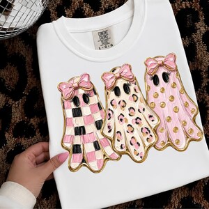 May include: A white t-shirt featuring a graphic of three ghosts with pink bows. The first ghost has a pink and black checkerboard pattern, the second has a leopard print, and the third has gold polka dots. The Comfort Colors label shows size L G G.