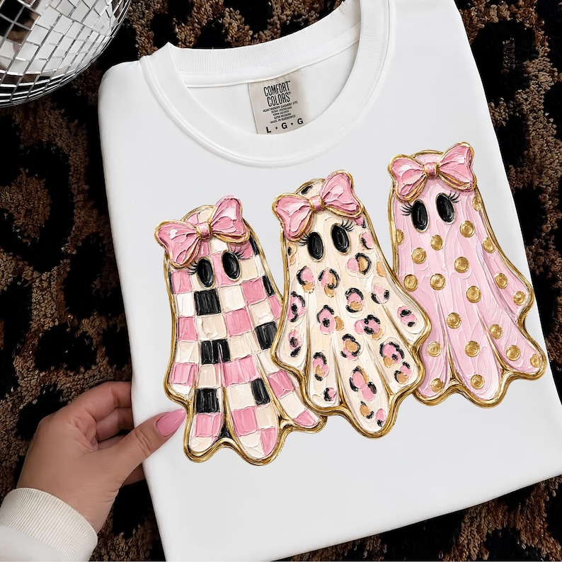 May include: A white t-shirt featuring a graphic of three ghosts with pink bows. The first ghost has a pink and black checkerboard pattern, the second has a leopard print, and the third has gold polka dots. The Comfort Colors label shows size L G G.