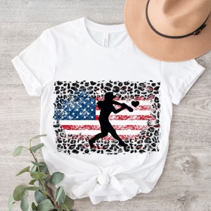 May include: White t-shirt with a graphic of a baseball player silhouetted against an American flag with a leopard print background.