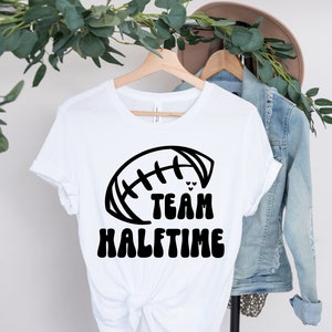 May include: White t-shirt with a black graphic of a football and the text "TEAM HALFTIME".
