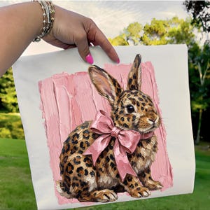 May include: A canvas print featuring a detailed illustration of a rabbit with a leopard print pattern. The rabbit has a large pink bow and is set against a textured pink background. The artwork is held up by a hand with pink nail polish.