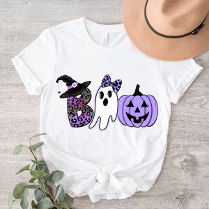 May include: White t-shirt featuring a purple, black and green "BOO" graphic. The "B" is a witch's hat with a spider, the "O" is a ghost with a bow, and the "O" is a purple pumpkin with a jack-o'-lantern face.