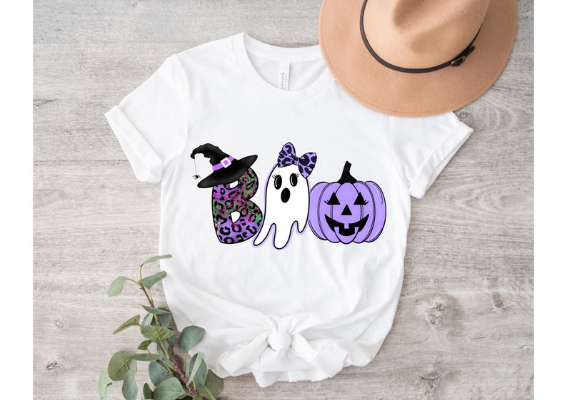 May include: White t-shirt featuring a purple, black and green "BOO" graphic. The "B" is a witch's hat with a spider, the "O" is a ghost with a bow, and the "O" is a purple pumpkin with a jack-o'-lantern face.
