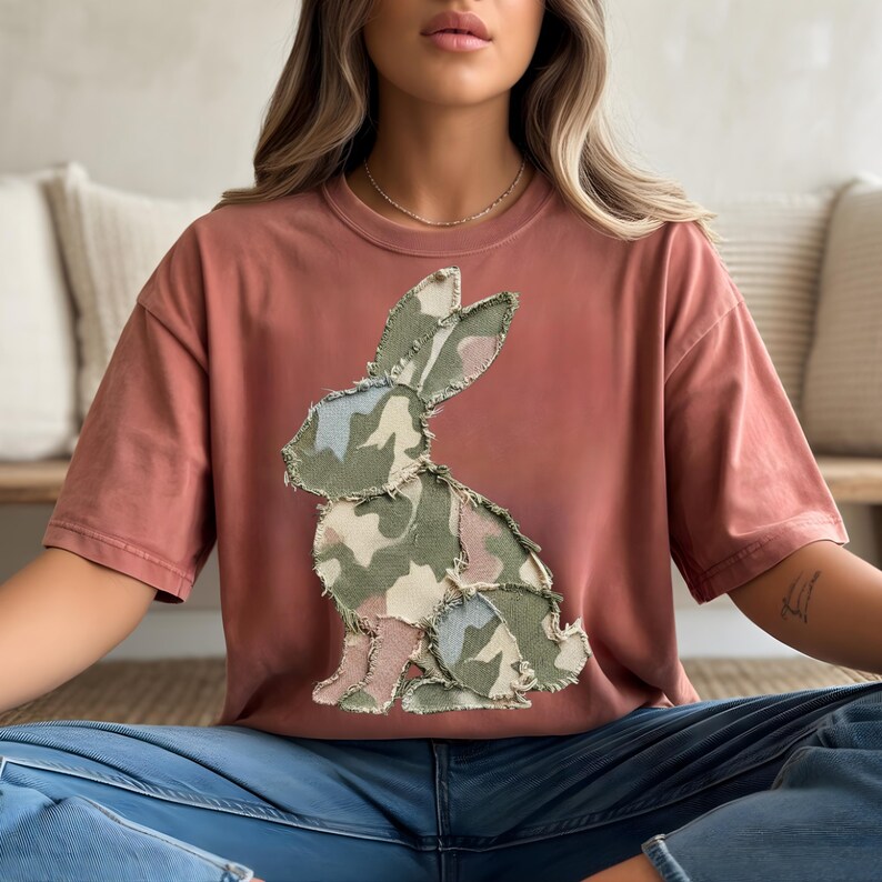 May include: A terracotta-coloured t-shirt with a camouflage bunny applique. The bunny design incorporates shades of green, pink, and blue. The shirt has short sleeves and a relaxed fit, suitable for casual wear.
