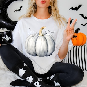 May include: A white t-shirt with a white pumpkin graphic and black and white swirls. The pumpkin has a gold stem and is surrounded by black dots. The shirt is being worn by a person with long blonde hair.