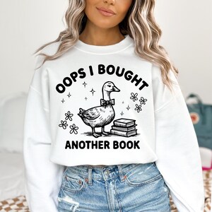 May include: White sweatshirt with a black and white graphic of a goose wearing a bow tie, with the text "OOPS I BOUGHT ANOTHER BOOK". The design includes stars and floral accents, perfect for book lovers.