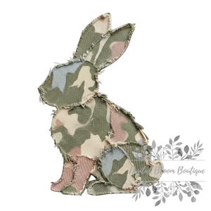 May include: A fabric bunny silhouette in a camouflage pattern of green, pink, and beige. The edges are frayed, giving a rustic appearance. The bunny is sitting, with its ears perked up.