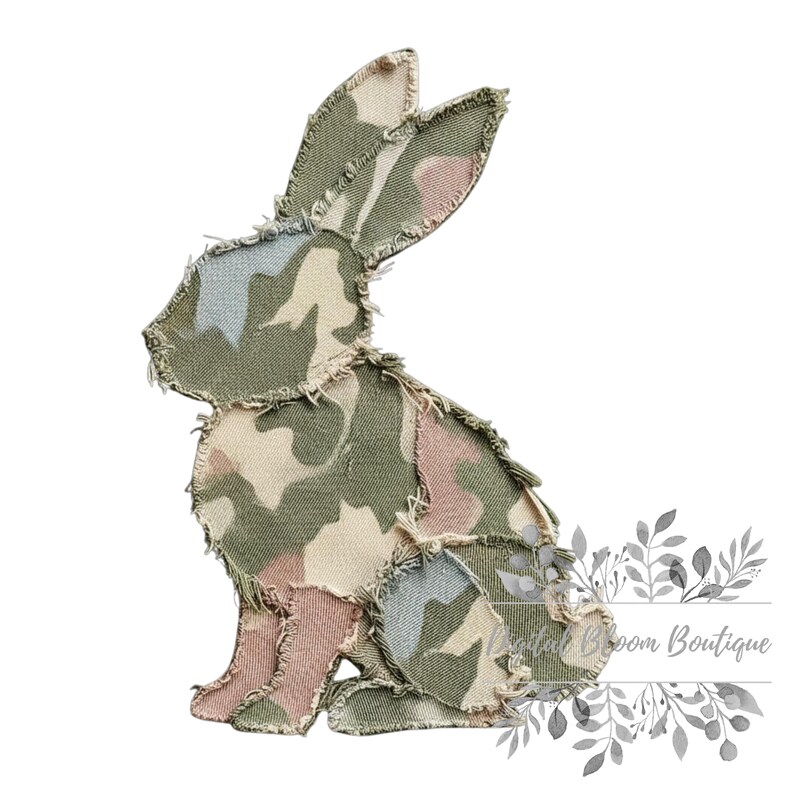 May include: A fabric bunny silhouette in a camouflage pattern of green, pink, and beige. The edges are frayed, giving a rustic appearance. The bunny is sitting, with its ears perked up.