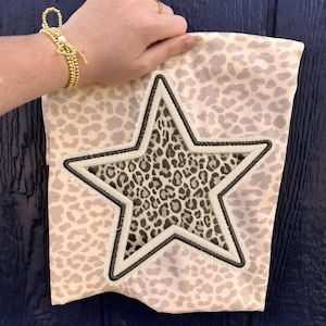 May include: A light pink fabric bag with a leopard print pattern. A large star with a leopard print center and a cream-colored border is embroidered on the front. The star has a dark brown outline and a fringed edge.