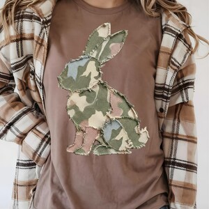 May include: A brown t-shirt with a camouflage bunny design. The bunny is made of green, pink, and blue fabric patches. The person is wearing a plaid shirt over the t-shirt.
