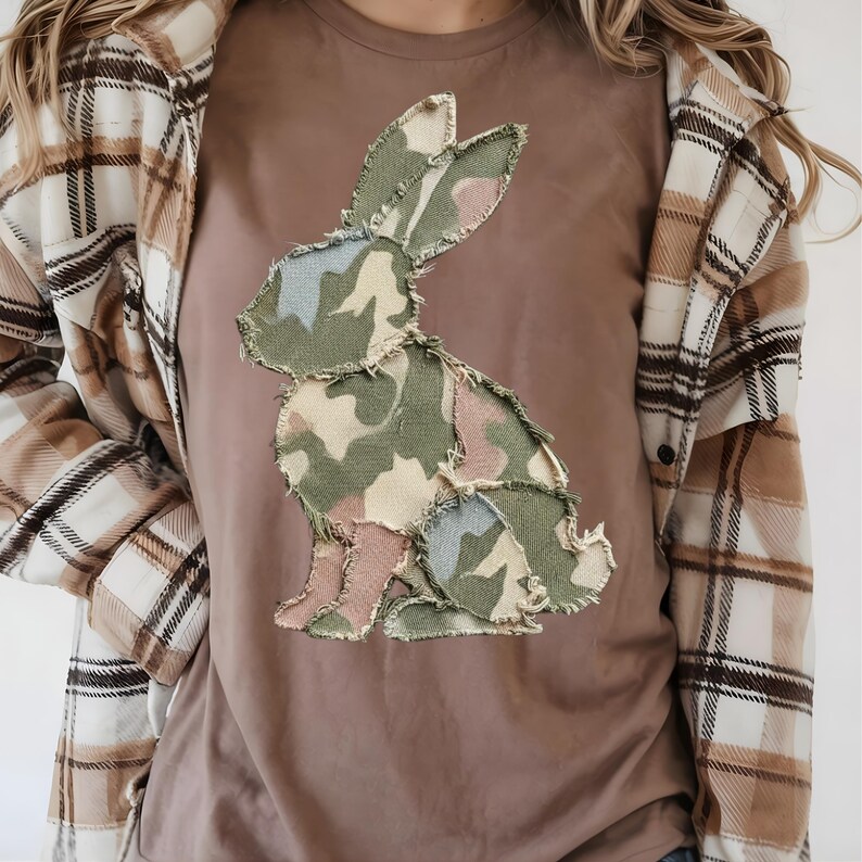 May include: A brown t-shirt with a camouflage bunny design. The bunny is made of green, pink, and blue fabric patches. The person is wearing a plaid shirt over the t-shirt.
