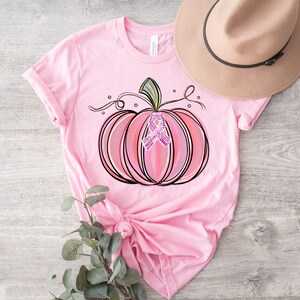 May include: Pink t-shirt featuring a pink and white pumpkin with a pink ribbon in the centre. The pumpkin has a green stem and vine.