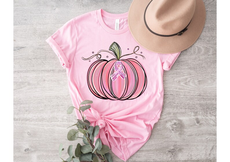 May include: Pink t-shirt featuring a pink and white pumpkin with a pink ribbon in the centre. The pumpkin has a green stem and vine.