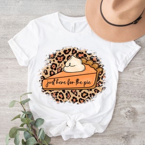 May include: White t-shirt with a graphic of a slice of pumpkin pie with whipped cream on top. The pie is on a leopard print background with the text "just here for the pie".
